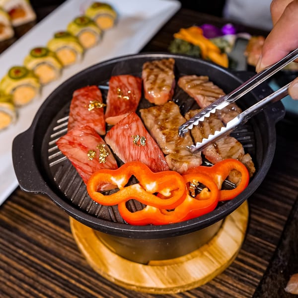 A hand uses tongs to flip wagyu beef and red bell peppers cooking on a grill pan.