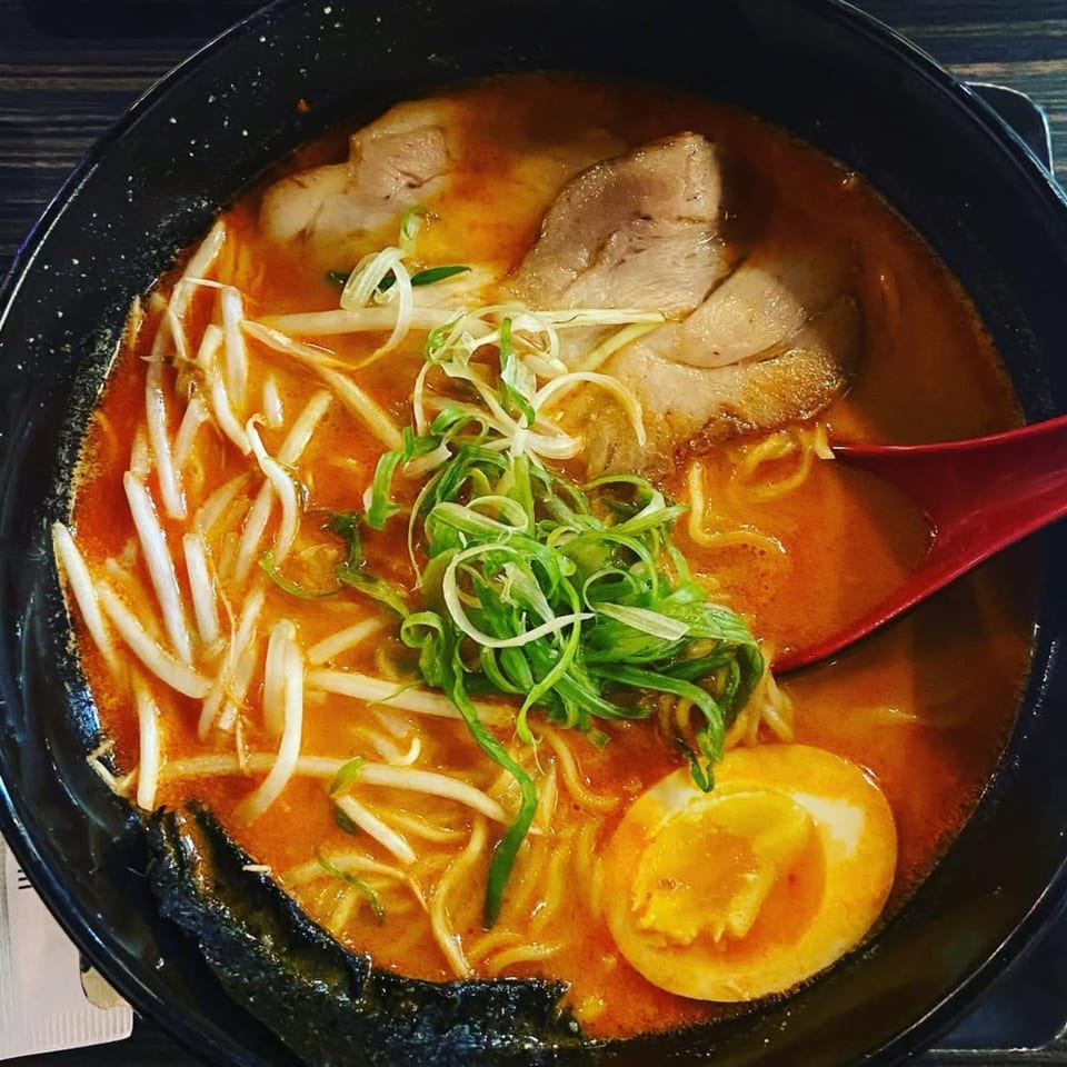 Spicy ramen with pork, bean sprouts, green onions, and a soft-boiled egg in a black bowl.