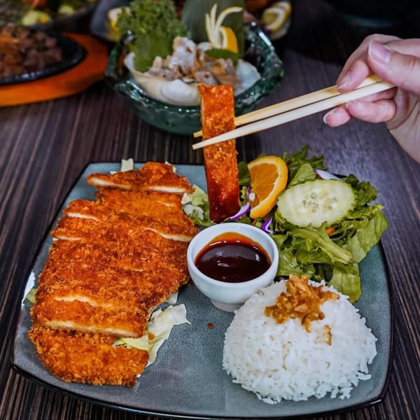 Chopsticks holding a piece of crispy fried chicken over a plate with rice, salad, and sauce.