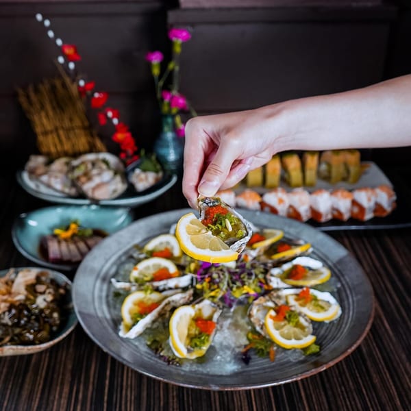 A hand picks up an oyster topped with lemon, roe, and green onions from a platter of oysters.