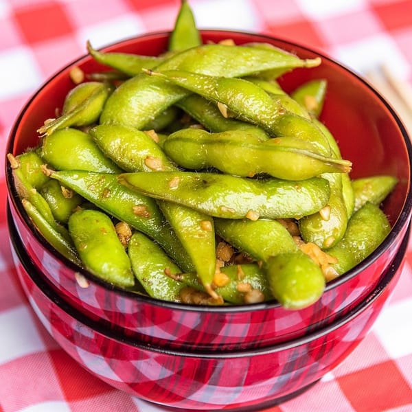 A red bowl filled with bright green edamame pods, sprinkled with garlic, on a red and white checkered tablecloth.
