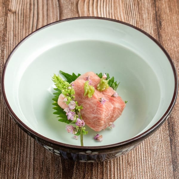 Wagyu beef sashimi with wasabi, shiso leaf, and edible flowers in a light green bowl.