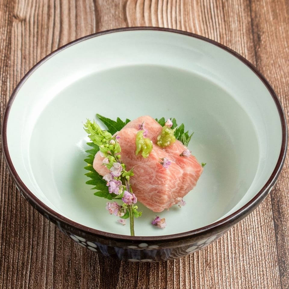 Wagyu beef sashimi with wasabi, shiso leaf, and edible flowers in a light green bowl.
