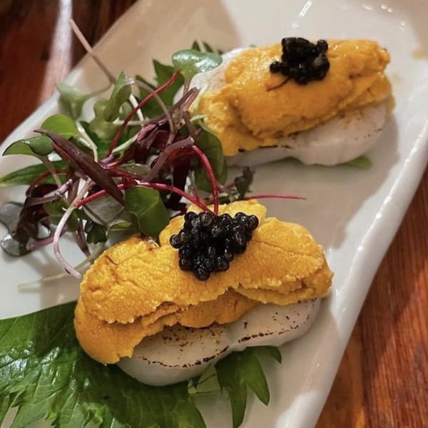 Two pieces of uni and scallop sushi topped with black caviar and microgreens on a white plate.