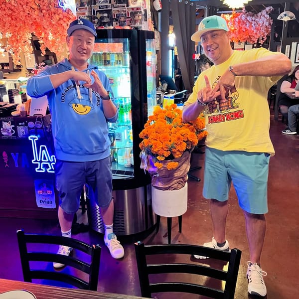 Two men in casual attire pose with hand gestures inside Sushiya, featuring a neon LA sign and cherry blossoms.