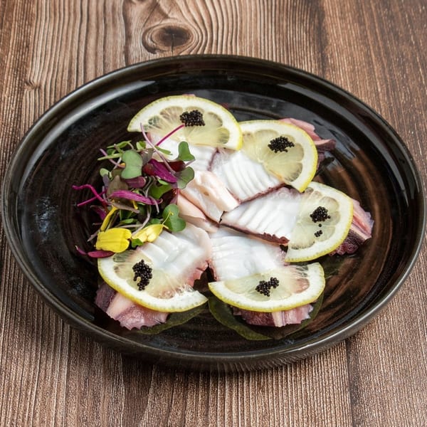 Octopus carpaccio with lemon slices, caviar, and microgreens on a dark plate.