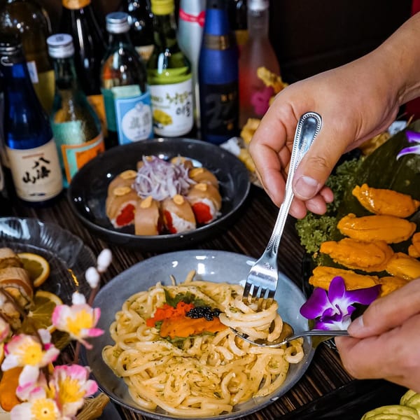 Hands twirling creamy udon noodles with uni and caviar, surrounded by sushi and sake bottles.