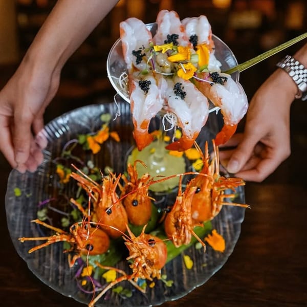 Hands presenting a martini glass of shrimp sashimi with caviar and edible flowers over a plate of shrimp heads.